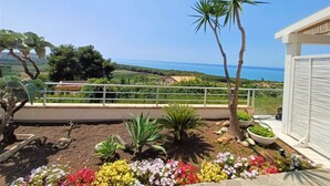 Jardí amb diverses plantes i flors, una palmera i vista al mar.