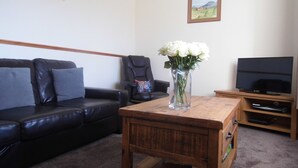 Un salon avec un canapé en cuir noir, une table basse en bois sur laquelle est posé un vase de fleurs blanches, un fauteuil en cuir noir, un téléviseur sur un meuble en bois et un tableau encadré accroché au mur.