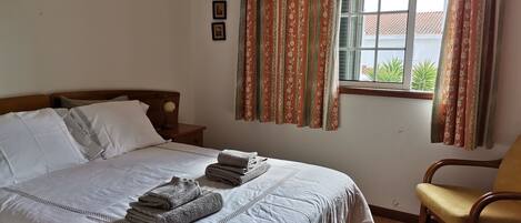 A neatly made bed with pillows and folded towels, a wooden nightstand with a lamp, a wooden chair, and a window with curtains.