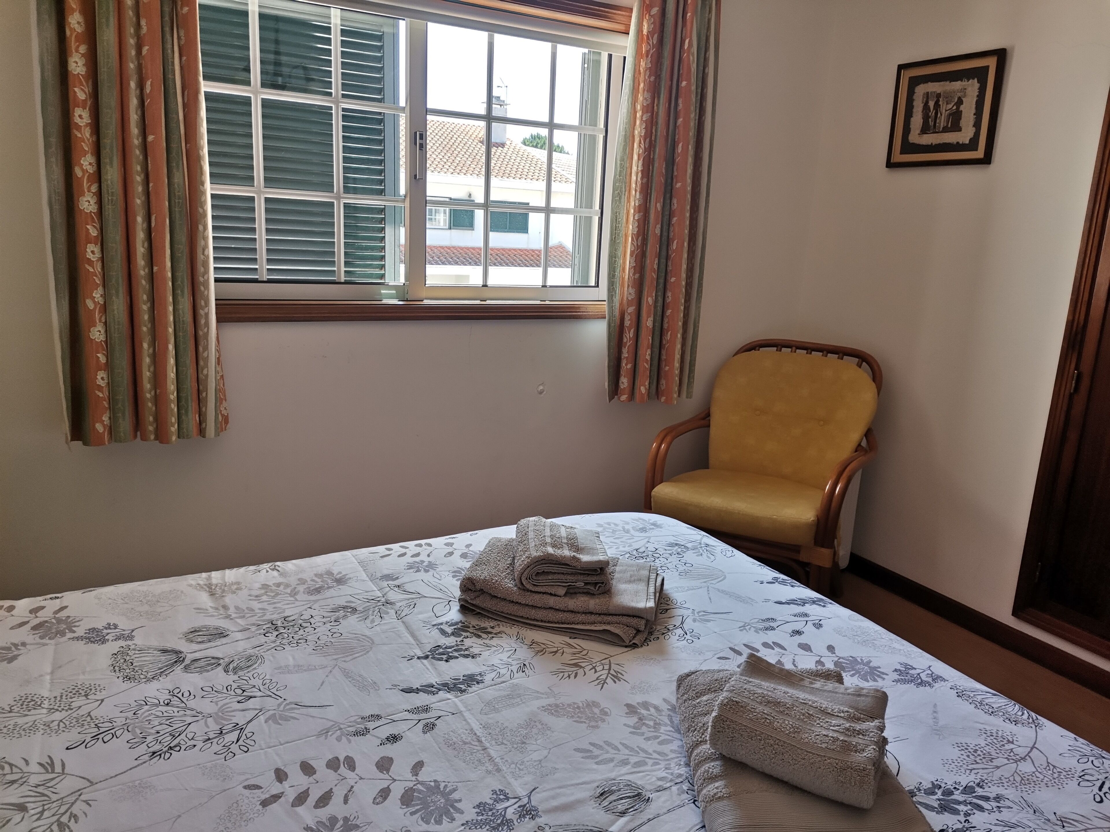 Un dormitorio con cama, una silla, un ventanal con cortinas y un cuadro en la pared.