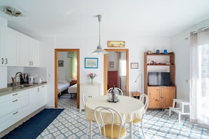 Une cuisine compacte avec des meubles blancs, une table à manger avec des chaises, un petit réfrigérateur et un téléviseur posé sur un meuble en bois.