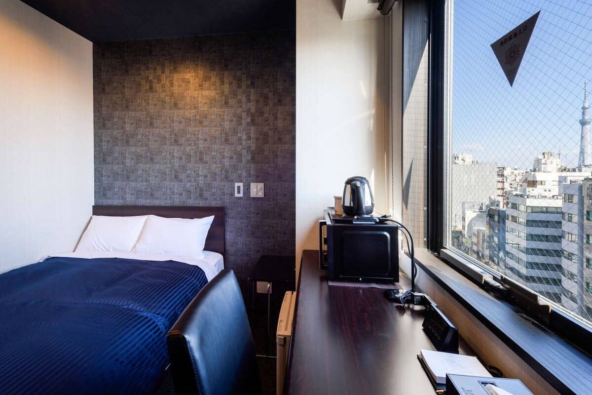 A hotel room with a bed, a desk with a kettle, and a view of the cityscape through a window.