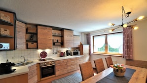 A modern kitchen with wooden cabinets, a dining table with chairs, a coffee maker, and a window with curtains.