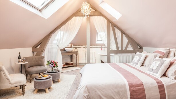 Een gezellige slaapkamer met een schuine kap, een bed met gestreepte beddengoed, een klein rond tafeltje, een stoel en een kaptafel met spiegel.
