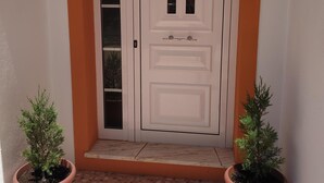 Une porte blanche ornée d’un panneau décoratif et d’une petite fenêtre, flanquée de deux plantes en pot sur un sol carrelé.