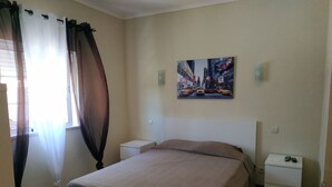Une chambre à coucher avec un lit, deux tables de chevet, une commode, une fenêtre avec des rideaux légers et un tableau représentant un paysage urbain accroché au mur.