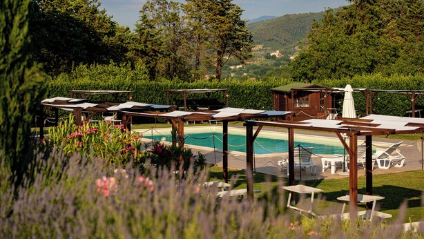 Zona junto a la piscina con sillones de descanso y sombrillas, rodeada de vegetación y con vistas a un pueblo lejano.