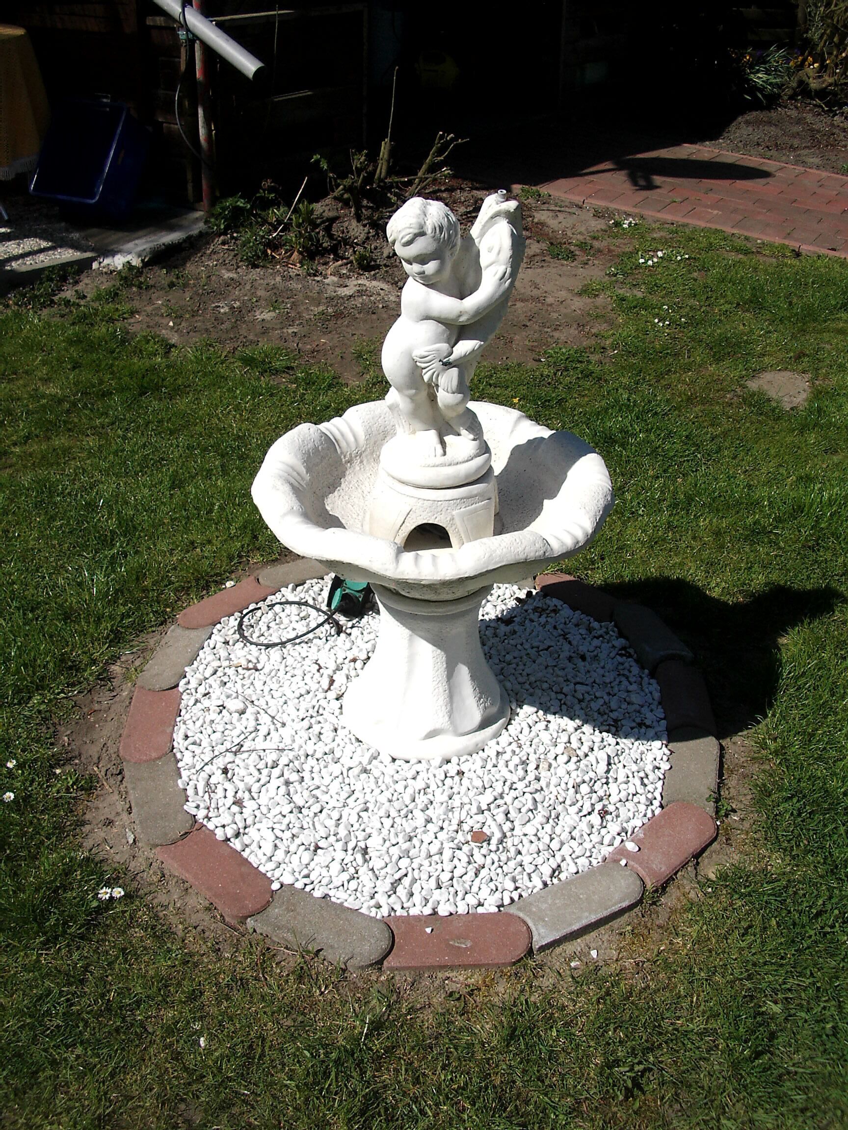Escultura blanca al aire libre de una figura con un ave, rodeada por un lecho circular de grava y un borde de ladrillo.