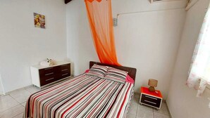 A bedroom with a bed, a nightstand, a bedside table, a wall-mounted air conditioner, and a window with curtains. - A rejuvenating stay in our peaceful gîtes (SAINTE ROSE)