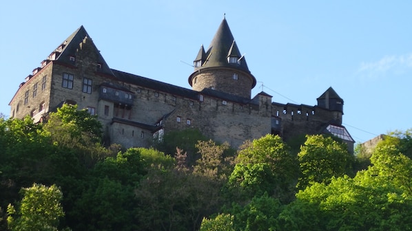 A large stone castle with multiple towers and a prominent round tower, surrounded by lush greenery.