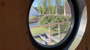 A round window with a metal railing, overlooking a grassy area with trees and a building in the background.