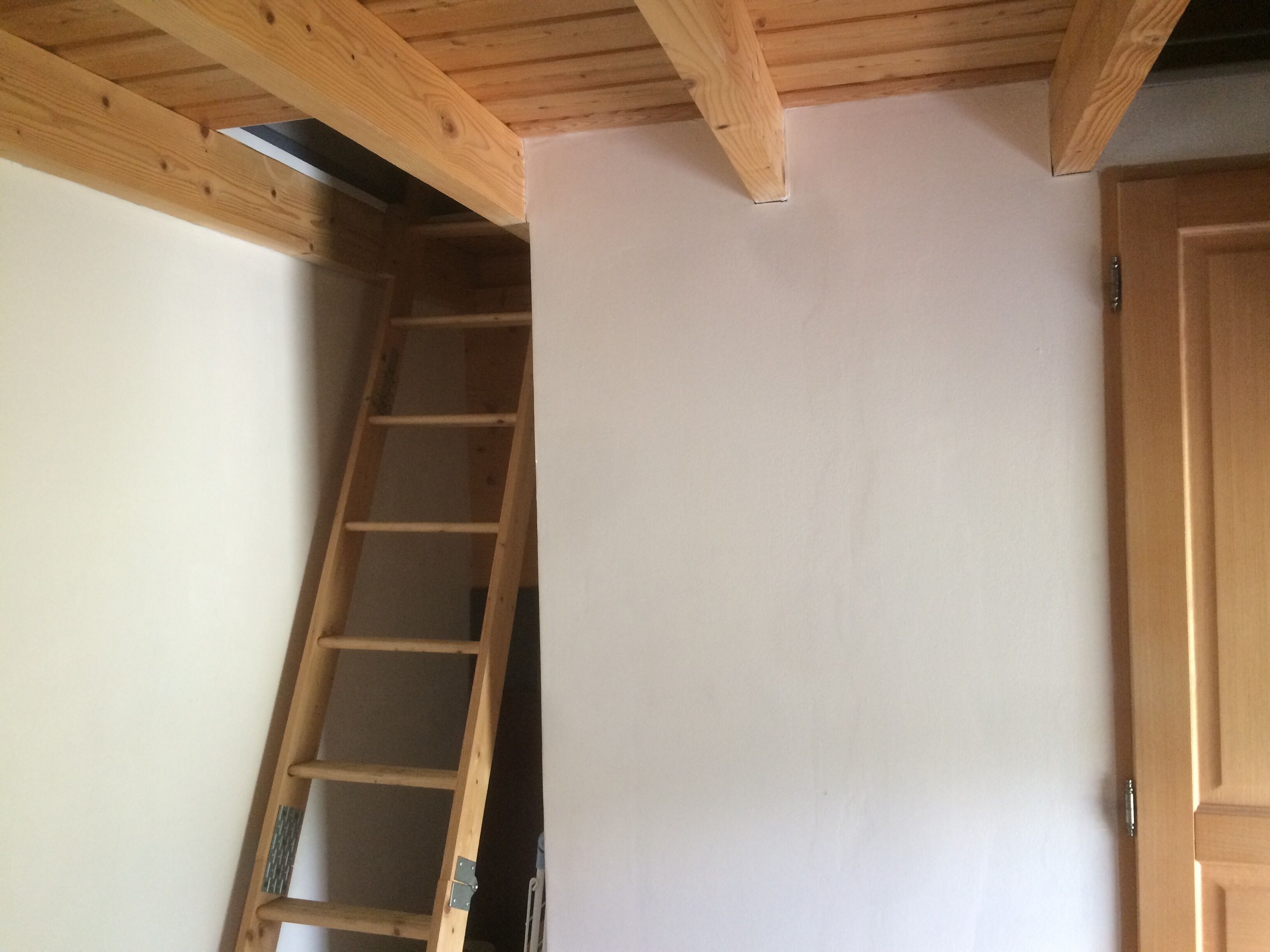 Ein Zimmer mit einer Holzleiter an einer weißen Wand, einer Holztür und einer geneigten Decke mit freiliegenden Balken.