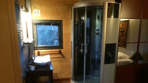 A room with a wooden floor, a small desk with a mirror, a window, and a glass-enclosed shower area.
