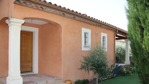 A two-story house with a peach-colored exterior, a wooden door, and a small garden with a tree and a bush.
