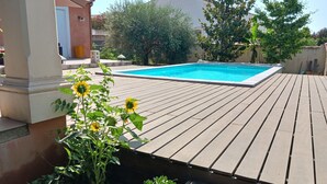 A wooden deck with a swimming pool, surrounded by greenery and a building in the background.