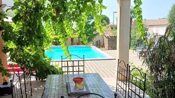 A covered patio with a table and chairs, a pool in the background, and a view of the surrounding area.