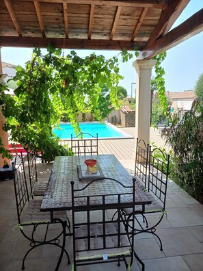 Un patio techado con mesa y sillas, una piscina al fondo y vistas del entorno.