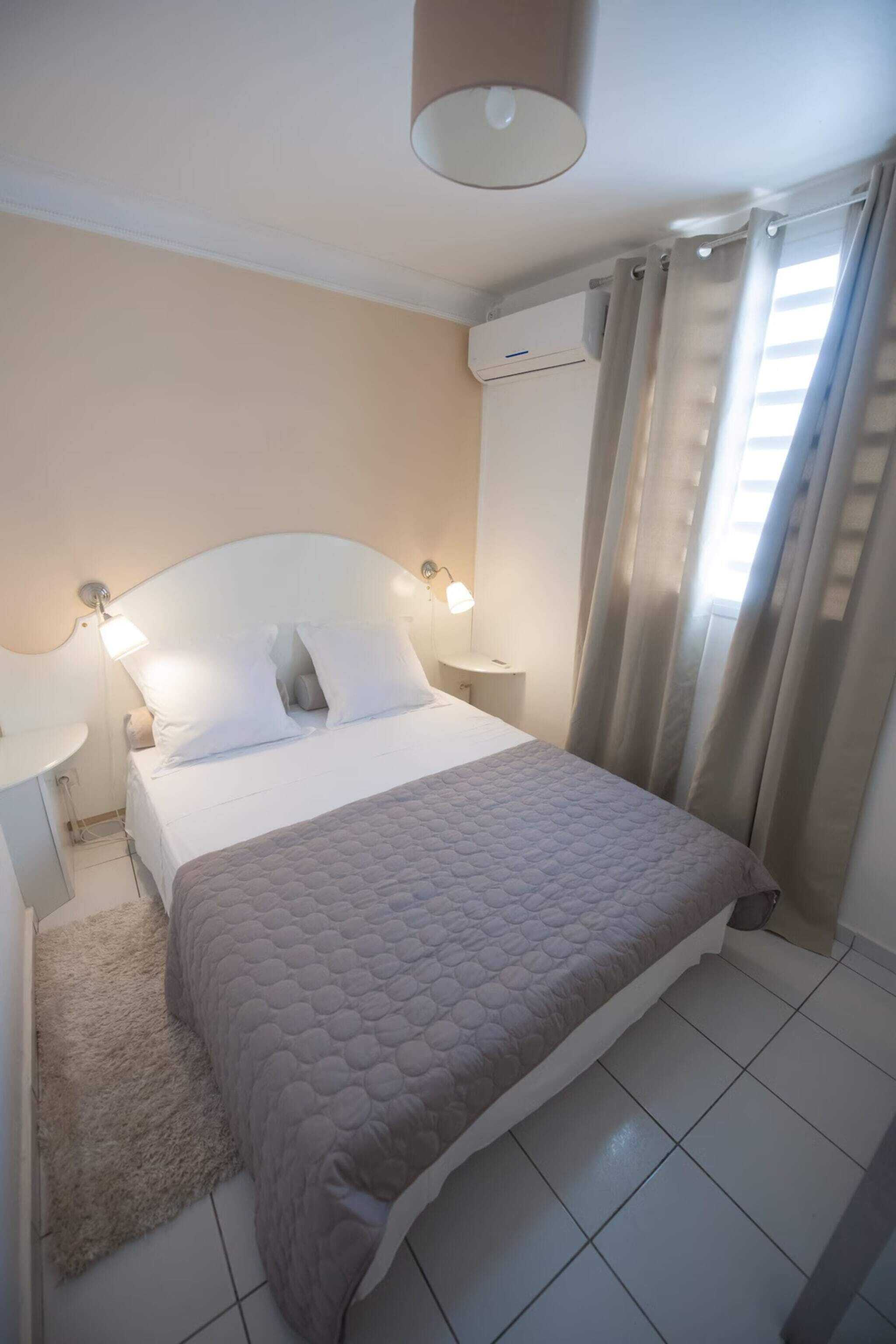 Un dormitorio con cama, lámparas de noche, una mesita, un tapete y un equipo de aire acondicionado.