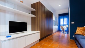 A modern living room with a flat-screen TV on a white console, a wooden cabinet, a blue sofa, and a dining area with a chair and table. - COZY FIRST FLOOR IN THE HEART OF ORTIGIA (Siracusa)