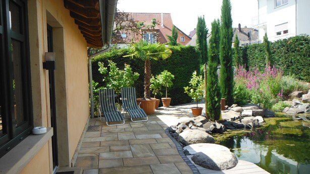 A patio with stone flooring, two striped lounge chairs, potted plants, a small pond, and a building in the background. - Guest house "Living in the garden of the senses" (Kindenheim)