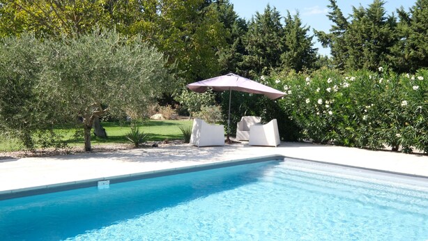 Una piscina con agua azul cristalina, rodeada por una terraza blanca. Hay dos sillones blancos y un paraguas morado.