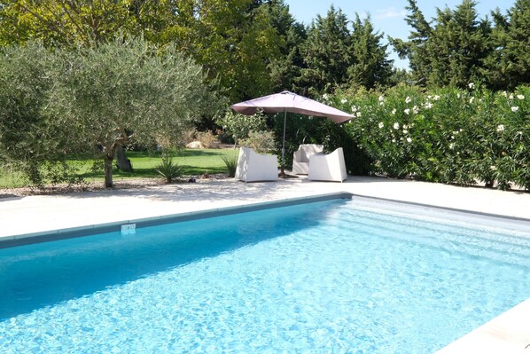 Une piscine à l’eau d’un bleu cristallin, entourée d’une terrasse blanche. On y trouve deux chaises longues blanches et un parasol violet.