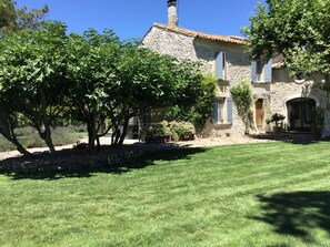Une maison en pierre avec un toit en tuiles, entourée d’une pelouse bien entretenue et d’arbres.