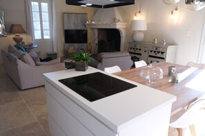 A modern kitchen with a central island, a dining area with a wooden table, and a living room with a sofa and a fireplace.