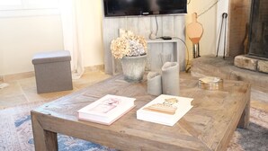 Una sala de estar con una mesa de centro de madera, un reposapiés gris, un jarrón con flores secas y una chimenea.