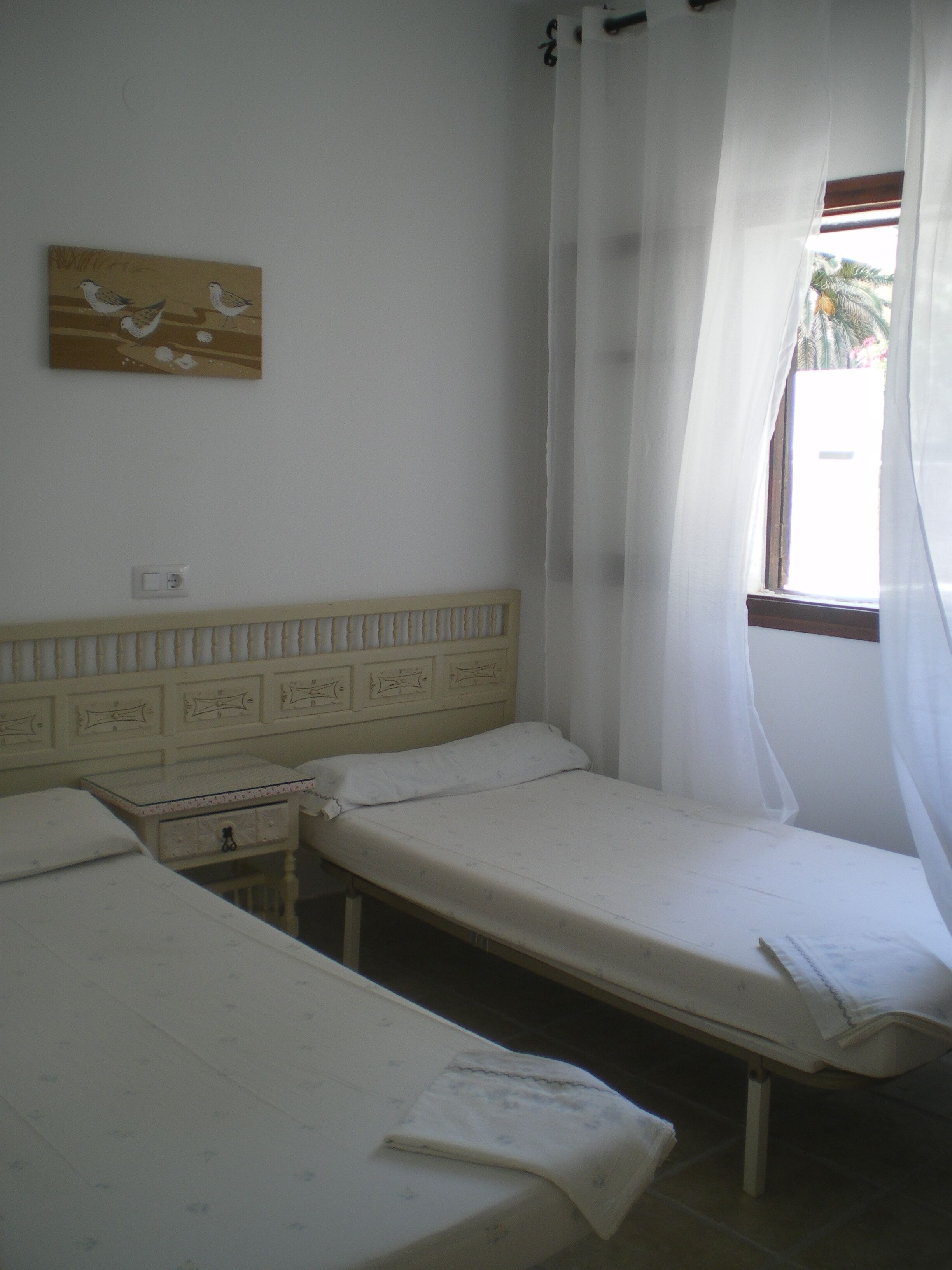 Une petite pièce simple, avec deux lits simples, une table de chevet, un interrupteur fixé au mur et une fenêtre ornée de rideaux légers.