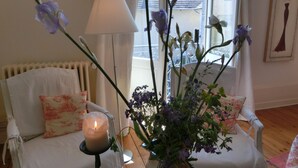 A room with a white armchair, a vase with flowers, a lit candle, and a window with a view of a building.