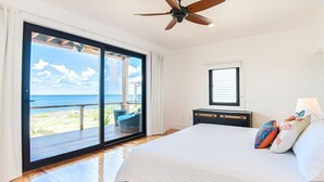 A bedroom with a large bed, a ceiling fan, a view of the ocean through a sliding glass door, and a nightstand with a lamp. - Vivaro South House Luxury Villa Private Infinity Pool (West End)