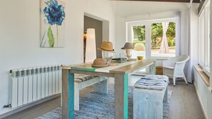 A cozy room with a wooden table, a bench, and a wicker chair. There's a large window with a view of greenery outside. - Beautiful villa with pool and garden,a/c, quiet location,  beach 200 mt (Cala Serena)