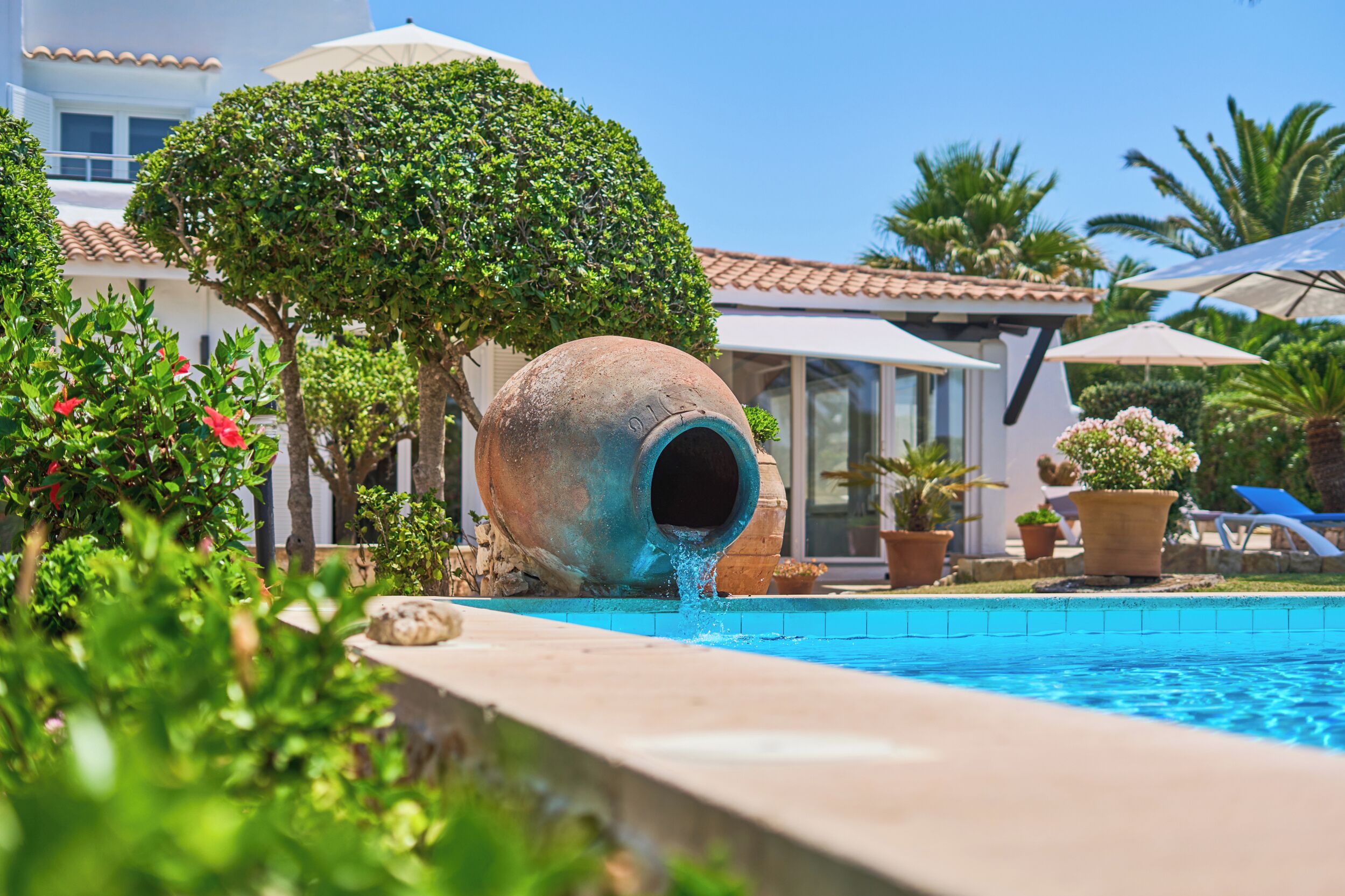 A swimming pool with a water feature, surrounded by lush greenery and a white building with a tiled roof.