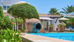 A swimming pool with a water feature, surrounded by lush greenery and a white building with a tiled roof. - Beautiful villa with pool and garden,a/c, quiet location,  beach 200 mt (Cala Serena)