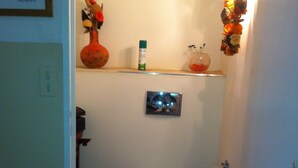 A small bathroom with a toilet, a shelf with a vase and a bottle, and a wall-mounted light fixture.