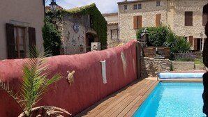 A pool area with a wooden deck, a red wall, and a view of traditional stone buildings in the background.