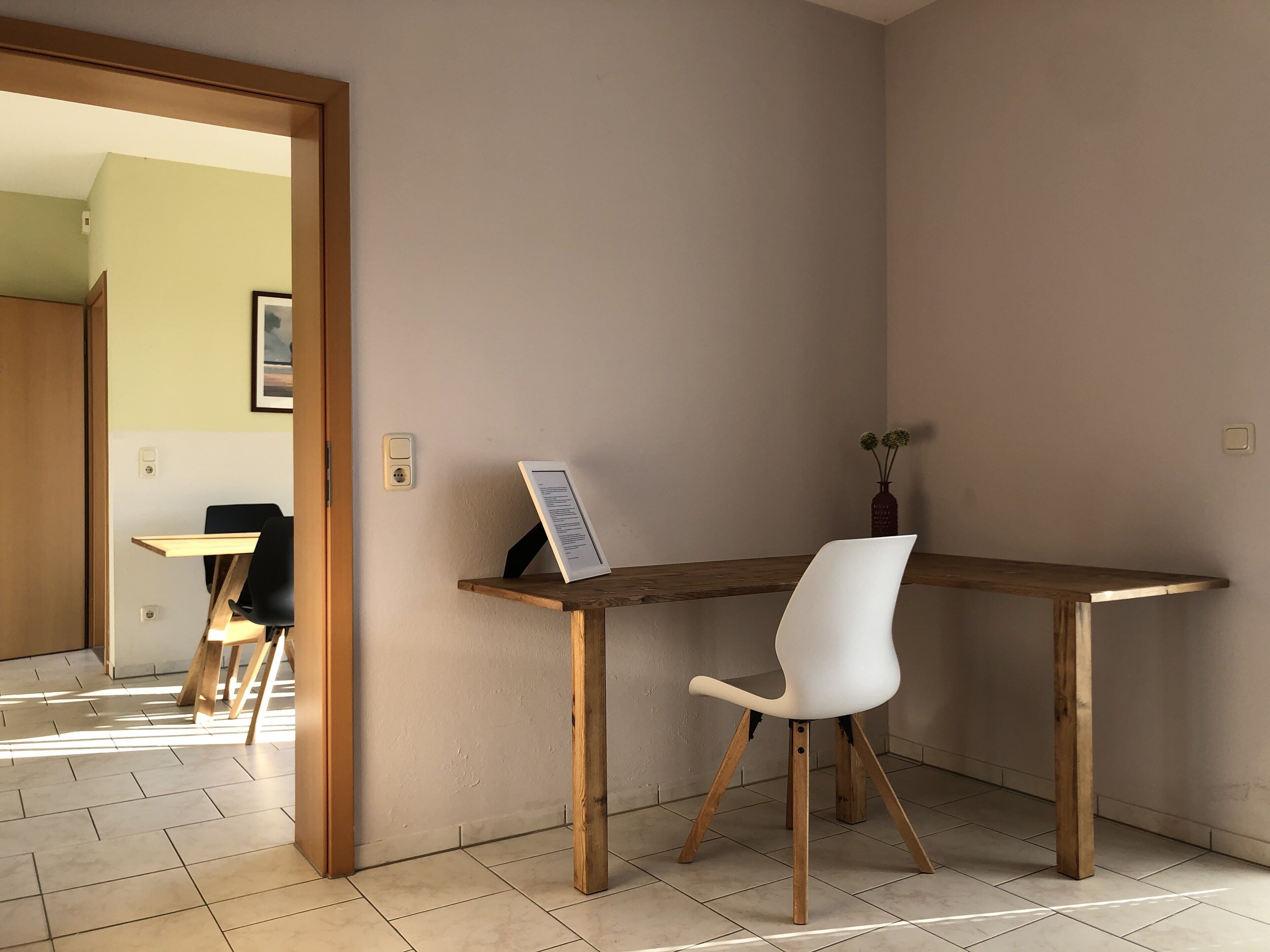 A room with a wooden desk, a white chair, a vase with flowers, and a framed picture on the wall.