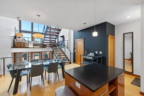 A modern kitchen with a central island, a dining area with a glass table, and a staircase leading to an upper level. - River Views! Contemporary Condo Centrally Located Walk To Restaurants/Breweries (Hood River)