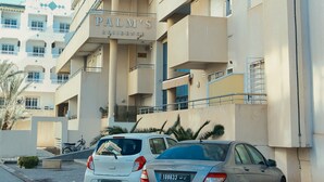 Un immeuble à plusieurs étages doté de balcons, une enseigne indiquant « RÉSIDENCE PALM’S », et deux voitures garées.