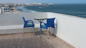 Un balcon avec deux chaises bleues et une petite table, donnant sur une plage et l’océan.