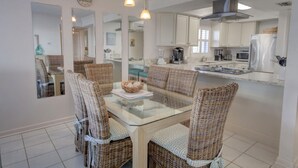 Cocina moderna con armarios blancos, una mesa de comedor de vidrio, sillas de mimbre y una isla central con cafetera.