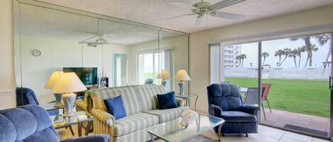 A living room with a sofa, armchairs, a coffee table, and a TV. There is a large mirror reflecting the room and a view of the outside.