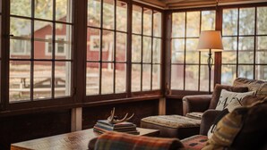 A cozy room with large windows, a leather sofa, a table with books, and a lamp. - Relax in Bayview Cabin MW Chain, June Opening (Manitowish Waters)