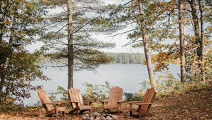 Three Adirondack chairs arranged around a small fire pit in a forested area near a lake. - Relax in Bayview Cabin MW Chain, June Opening (Manitowish Waters)