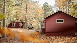 A red cabin with a small porch and a window, surrounded by trees with autumn leaves. - Relax in Bayview Cabin MW Chain, June Opening (Manitowish Waters)
