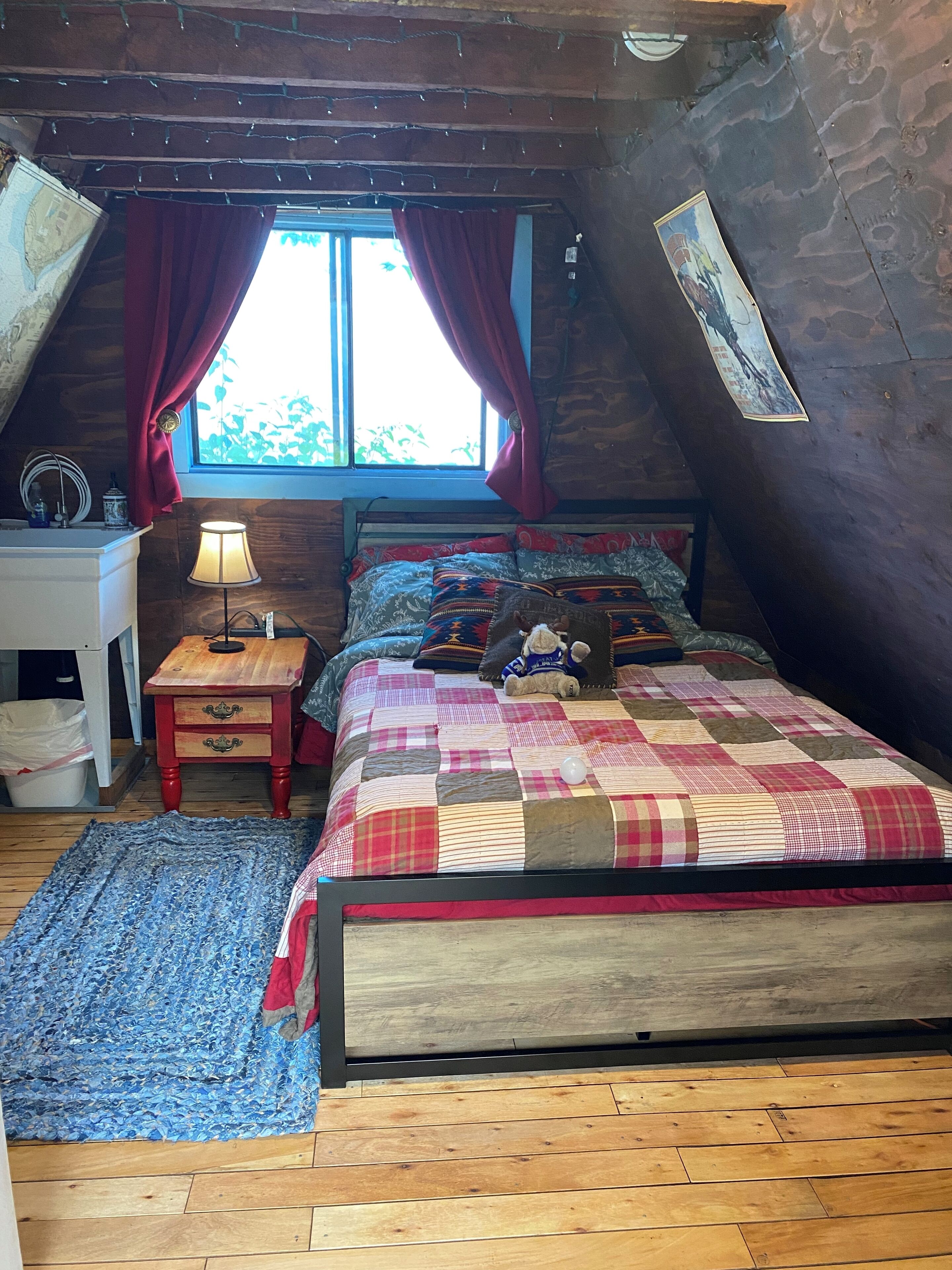 A cozy attic bedroom with a wooden floor, a bed with a quilt, a nightstand with a lamp, and a window with curtains.