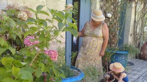 A woman in a floral dress and hat standing next to a dog, with large potted plants and a wooden deck in the background. - Claire s' house in medieval village couze near river Dordogne with your pets (Couze)