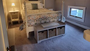 A bedroom with a four-poster bed, a nightstand with a lamp, a window with a view, and a storage bench with baskets. - The Nest - one bed apartment (Searsport)