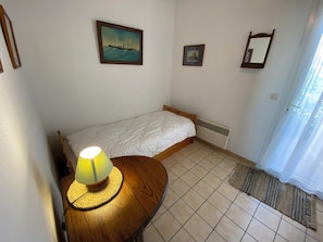 A small, tidy room with a single bed, a bedside table with a lamp, a framed picture on the wall, and a window with curtains.